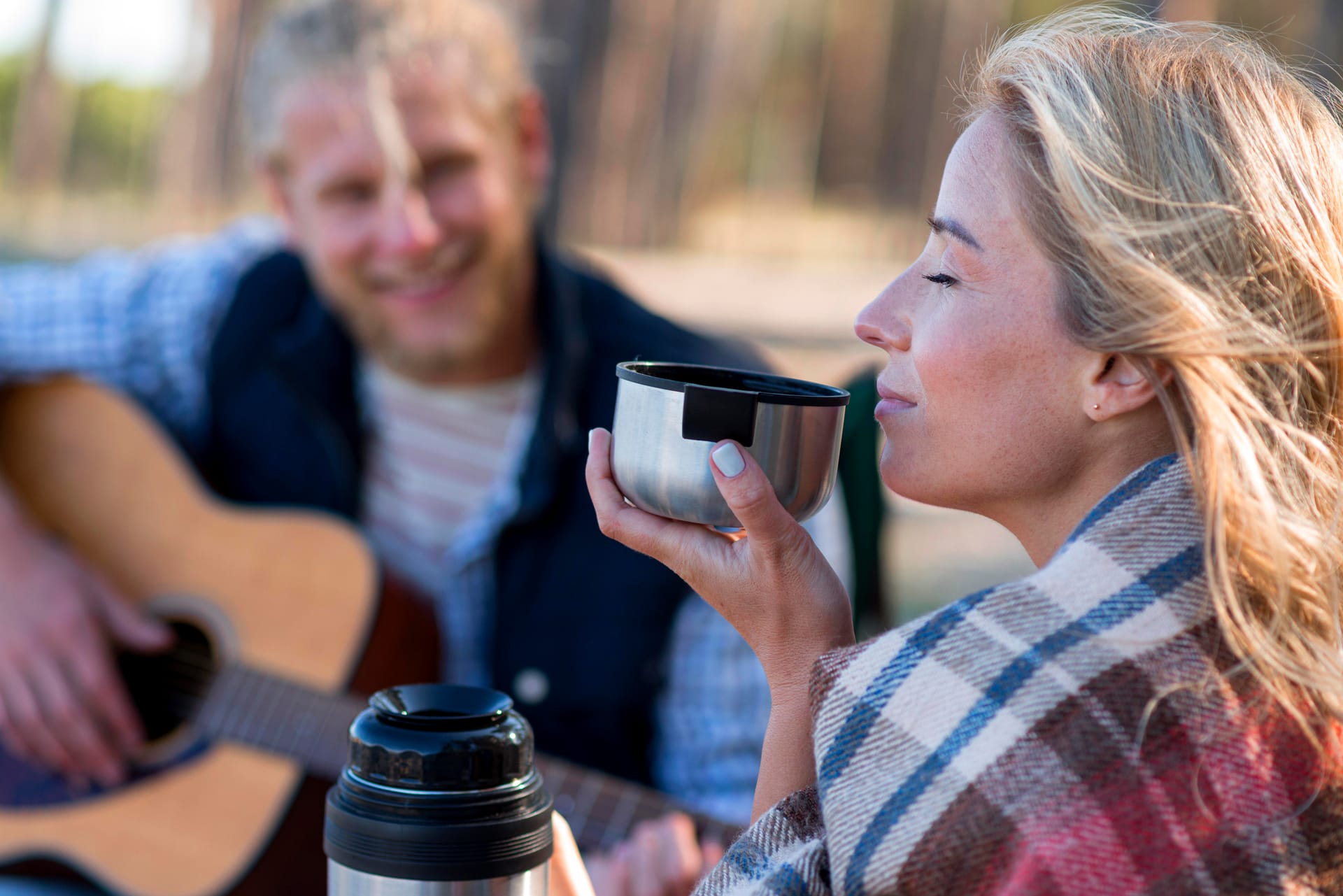 Woman with steaming coffee on a camping blanket beside a thermos and guitar
